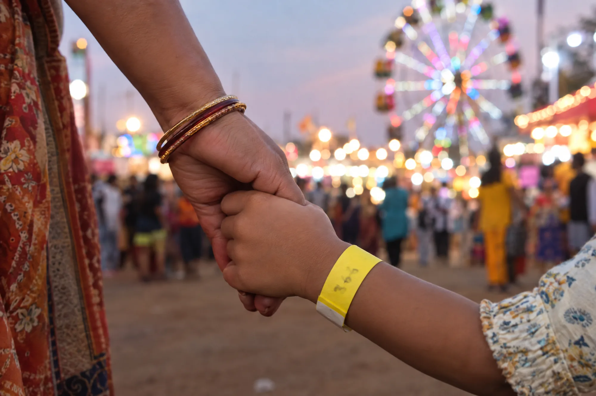A parent holding a child’s hand at an evening fair, the child wearing a yellow paper wristband with a number written on it in marker