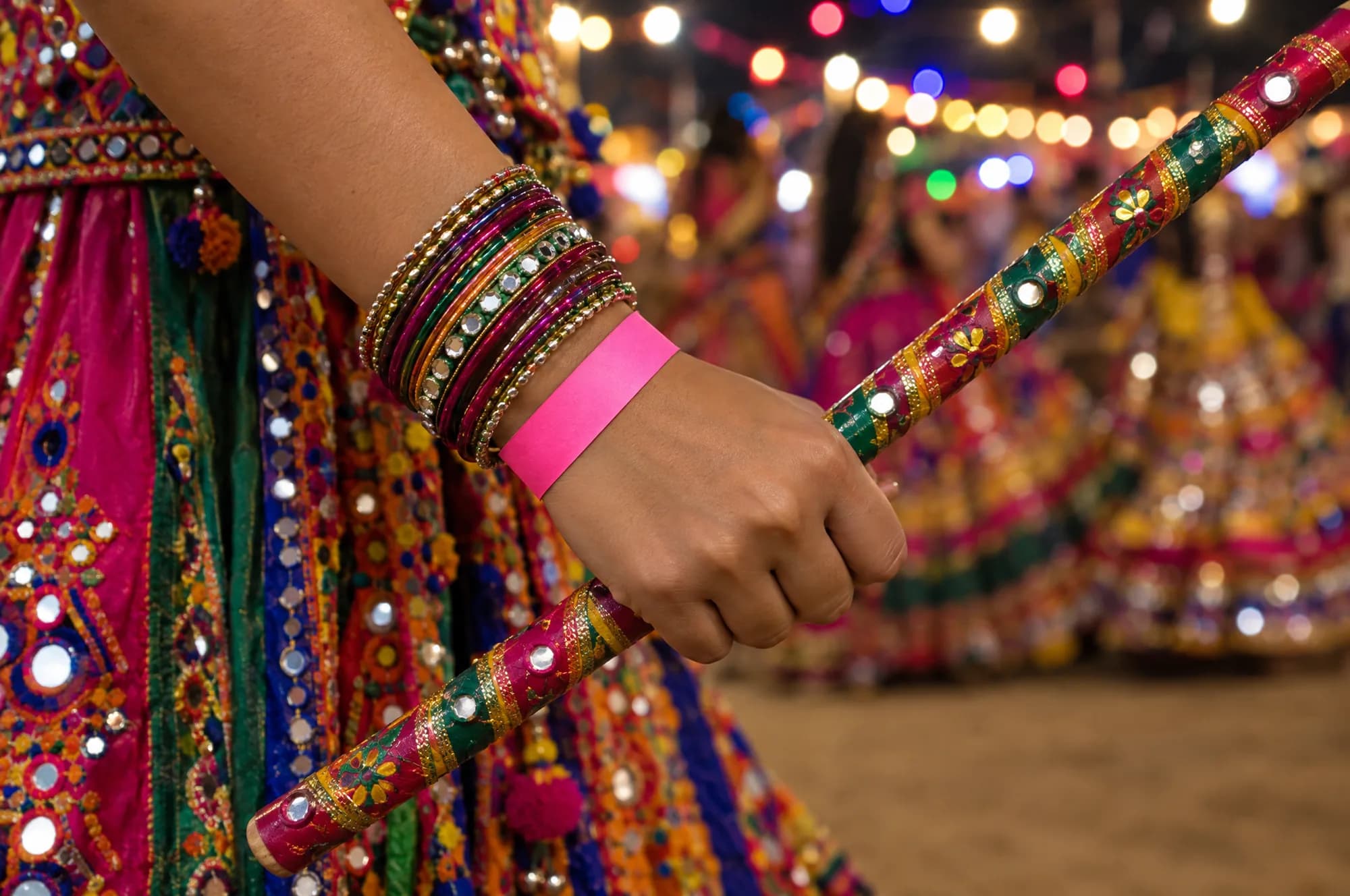 A Garba dancer’s hand with bangles and a dandiya stick, wearing a pink paper wristband