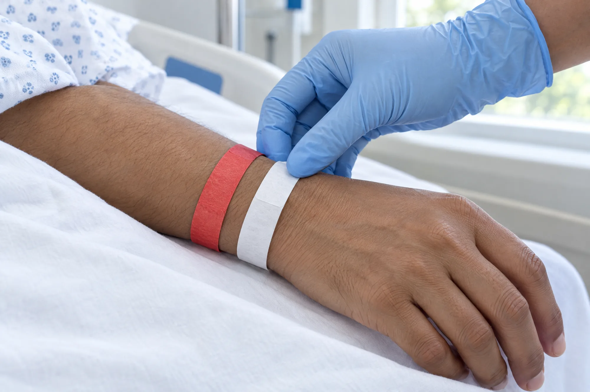 A nurse in a blue glove checking a white patient identification wristband on a patient’s wrist